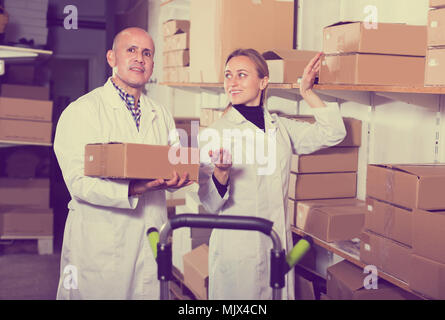 Smiling young female and male transporting cart cardboard cases in storage Stock Photo