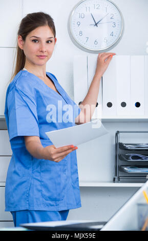 Smiling attractive female doctor searching patient history on shelves in office Stock Photo