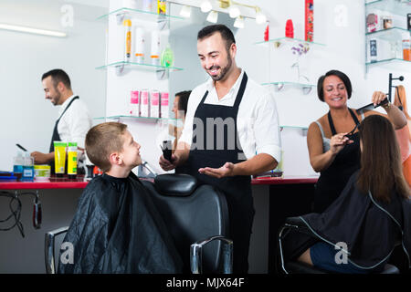 Smiling man hairdresser cutting hair boy in elementary school age in barber salon Stock Photo