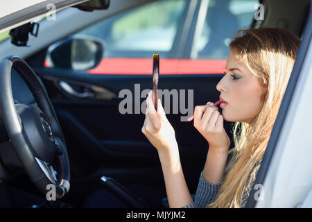 Young blonde woman applying lipstick looking at mirror in her car. Girl making-up herself in urban background. Stock Photo