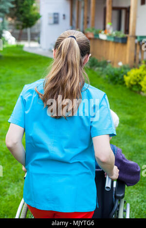 Caregiver spending time outdoor with handicapped senior woman, home care - back view Stock Photo