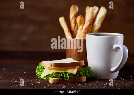 Breakfast table with fresh cheese and lettuce sandwich, breadsticks and black coffee in white ceramic mug on rustic wooden background, close-up, selec Stock Photo