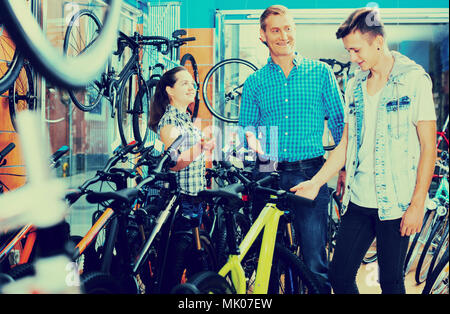 joyful smiling father with teenage son getting help from female seller in bike store. Focus on woman Stock Photo