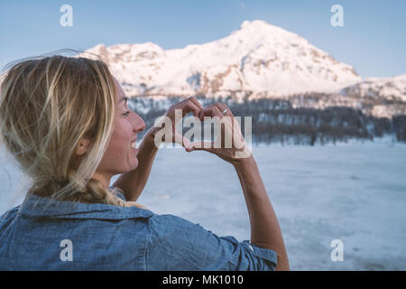 Young woman making a heart shape finger frame over frozen lake and snowcapped mountains. People love nature environment concept. Shot in Switzerland Stock Photo