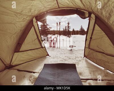 Camping in winter forest, cooking in front of tent in snow. Making a bonfire campfire and cooking food with portable gas cooker and fire, snowy landsc Stock Photo
