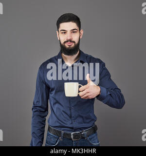 Handsome bearded man with stylish hair beard and mustache on serious face in shirt holding white cup or mug drinking tea or coffee in studio on grey background Stock Photo