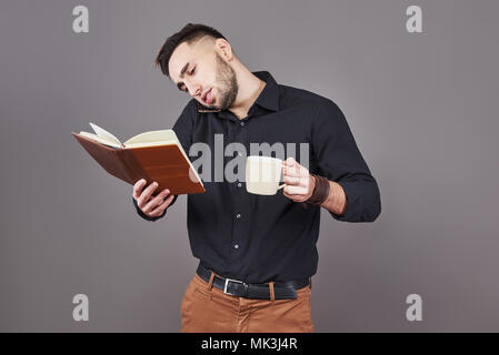 Portrait of young handsome man with phone, cup of coffe and book on hands. The concept of overtime, stress and a lot of work Stock Photo