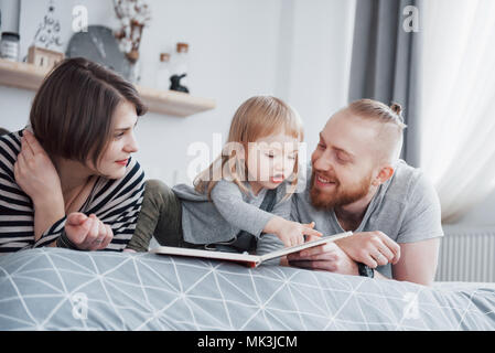 Father, Mother and Little Daughter Reading Children's Book on a Sofa in the Living Room. Happy big family read an interesting book on a festive day. Parents love their children Stock Photo