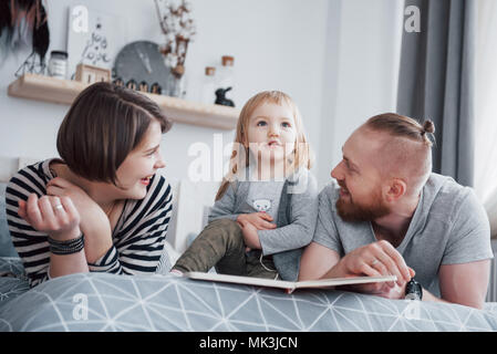 Father, Mother and Little Daughter Reading Children's Book on a Sofa in the Living Room. Happy big family read an interesting book on a festive day. Parents love their children Stock Photo