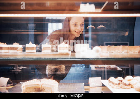 Smiling woman at camera through the showcase with sweet and cakes in modern cafe interior Stock Photo