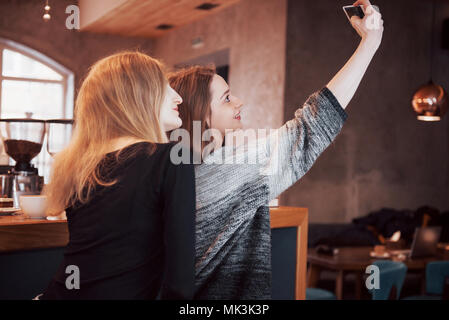 Two friends drinking coffee in a cafe, taking selfies with a smart phone and having fun making funny faces. Focus on the girl on the left Stock Photo