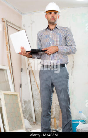 Smiling man engineer managing process with laptop in helmet indoors Stock Photo