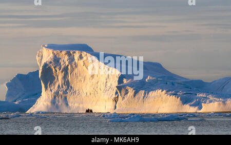 A zodiac full of tourist viewed against a large iceberg with the light of the midnight sun on the summer solstice, Cierva Cove, Antarctica Stock Photo
