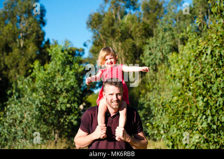 A young girl sits on her fathers shoulders in a park Stock Photo