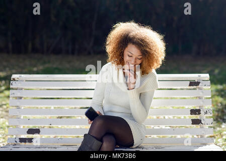 Beautiful young African American woman with afro hairstyle wearing white winter dress. Beautiful Girl sitting in the park with a smartphone. Stock Photo