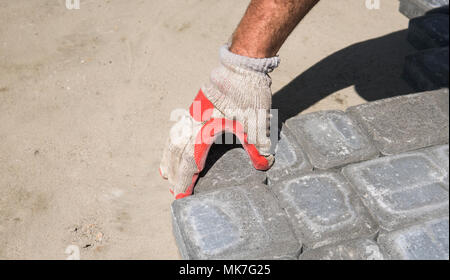Hands of a builder in his orange gloved hands fitting laying new exterior paving stones carefully placing one in position on a leveled and raked soil base. Sand foundation. Building. Stock Photo