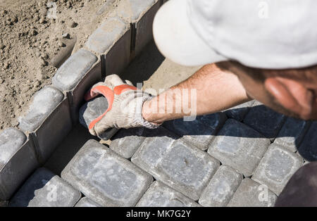 Hands of a builder in his orange gloved hands fitting laying new exterior paving stones carefully placing one in position on a leveled and raked soil base. Sand foundation. Building. Stock Photo