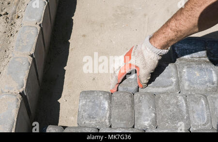 Hands of a builder in his orange gloved hands fitting laying new exterior paving stones carefully placing one in position on a leveled and raked soil base. Sand foundation. Building. Stock Photo
