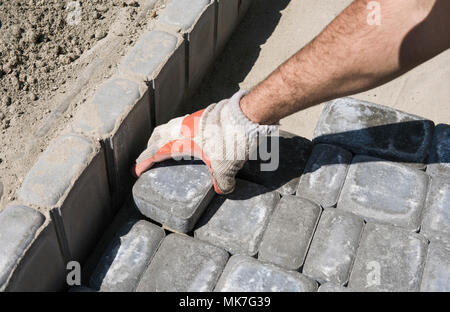 Hands of a builder in his orange gloved hands fitting laying new exterior paving stones carefully placing one in position on a leveled and raked soil base. Sand foundation. Building. Stock Photo