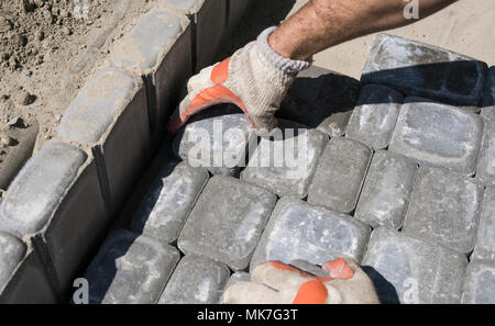 Hands of a builder in his orange gloved hands fitting laying new exterior paving stones carefully placing one in position on a leveled and raked soil base. Sand foundation. Building. Stock Photo