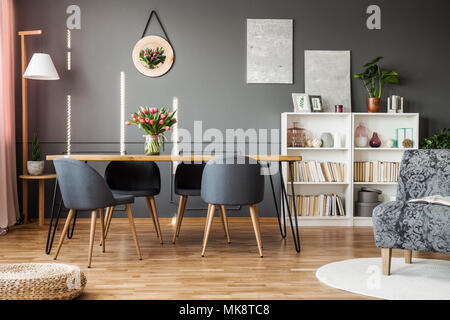 Grey chairs at wooden table with red tulips in dining room interior with books on shelves Stock Photo