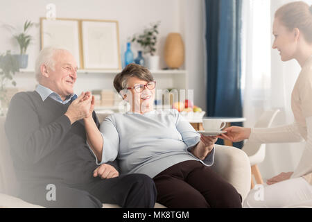 Young at heart emotional senior couple holding hands while sitting on settee and being served by a caregiver a cup of tea in a sunny living room at ho Stock Photo