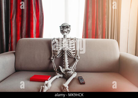 Smiling skeleton in glasses is sitting on couch between book and tv remote control, window and red curtains on background. Funny joke Stock Photo