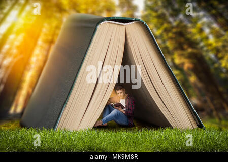 Reading concept, man sitting on the grass under large book and reads. Gaining knowledge from literature Stock Photo