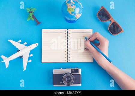 Female hand writing in blank notebook and airplane toy, palm tree, camera, sunglasses and globe model against blue background minimal creative concept Stock Photo