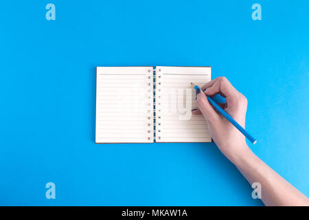 Female hand writing in blank notebook against blue background minimal creative concept. Space for copy. Stock Photo