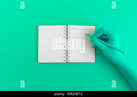 Female hand writing in blank notebook against green background minimal creative concept. Space for copy. Stock Photo