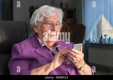 A 95 year old woman is holding a smartphone and is typing on the touch screen. She is sitting on a armchair in her living room and looks at the displa Stock Photo