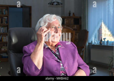A 95 year old woman is holding a smartphone to her ear. She is sitting on a armchair in her living room Stock Photo