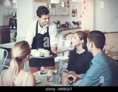 Polite smiling waiter bringing ordered dishes to friends in tearoom of a cozy confectioner's shop Stock Photo