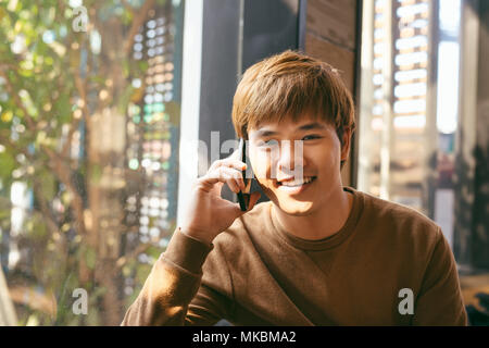 Closeup portrait of smiling young Asian man talking on mobile phone and sitting at empty table with blurred cafe interior in background Stock Photo