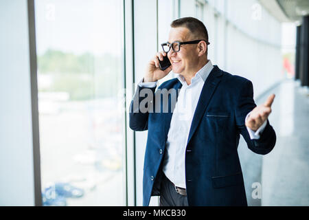 Positive confident successful businessman in formal suit standing at window and contemplating cityscape while talking on mobile phone on office Stock Photo