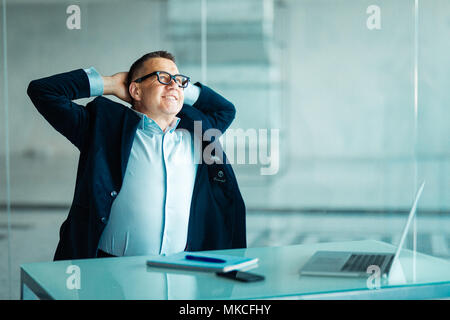 Successful businessman sitting pondering a problem or new idea relaxing back in his chair at the office with his hands behind his head Stock Photo