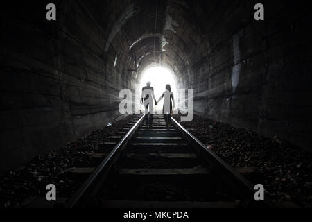 Couple walking hand in hand along the track through a railway tunnel towards the bright light at the other end, they appear as silhouettes against the Stock Photo