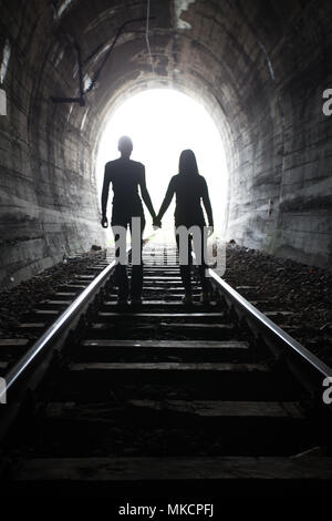 Couple walking hand in hand along the track through a railway tunnel towards the bright light at the other end, they appear as silhouettes against the Stock Photo