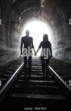 Couple walking hand in hand along the track through a railway tunnel towards the bright light at the other end, they appear as silhouettes against the Stock Photo