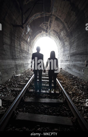 Couple walking hand in hand along the track through a railway tunnel towards the bright light at the other end, they appear as silhouettes against the Stock Photo