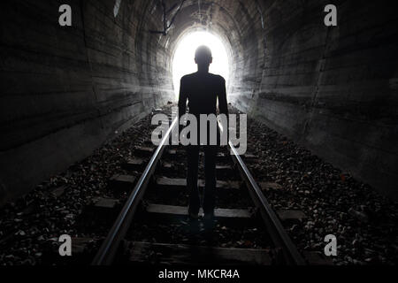 Man silhouetted in a tunnel standing in the center of the railway tracks looking towards the light at the end of the tunnel in a conceptual image Stock Photo