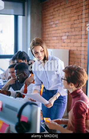 closeup portrait of two female executives working with paper in the loft office. Woman with long brown hair is giving documents to secretary in glasse Stock Photo