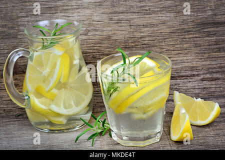 Lemon water with rosemary and ice cubes in a glass and pitcher on wood background Stock Photo