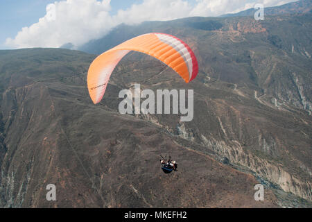 Huge orange paraglider with a man in a black suit hovers in the air against the background of a brown mountain slope. Stock Photo