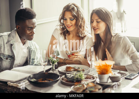 Three friends sitting at the served for lunch table with tasty delicious meal , european women and one african guy at cafe, watching photos on smartph Stock Photo