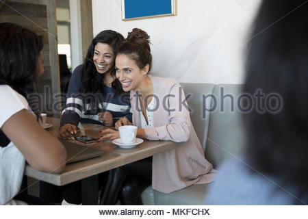 Smiling young women friends talking, drinking coffee and using smart phone in cafe