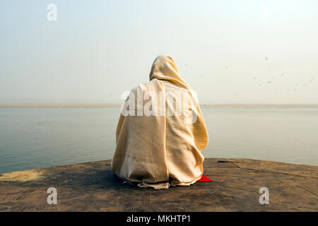 A holy man dressed in white is praying and meditating on one of the many Ghats of Varanasi in front of the sacred river Ganges, India. Stock Photo