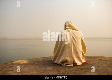 A holy man dressed in white is praying and meditating on one of the many Ghats of Varanasi in front of the sacred river Ganges, India. Stock Photo