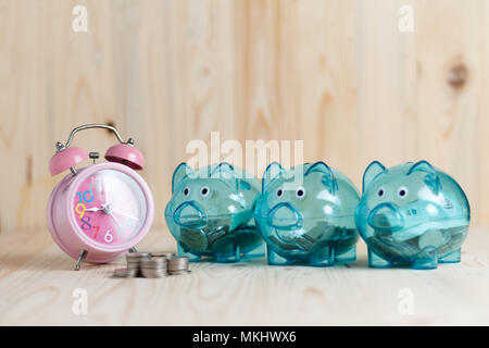 Alarm clock and coins put on the wood table. Clear piggy bank with 3 pigs, which has money in it. Placed on a wooden background. Saving money on time  Stock Photo